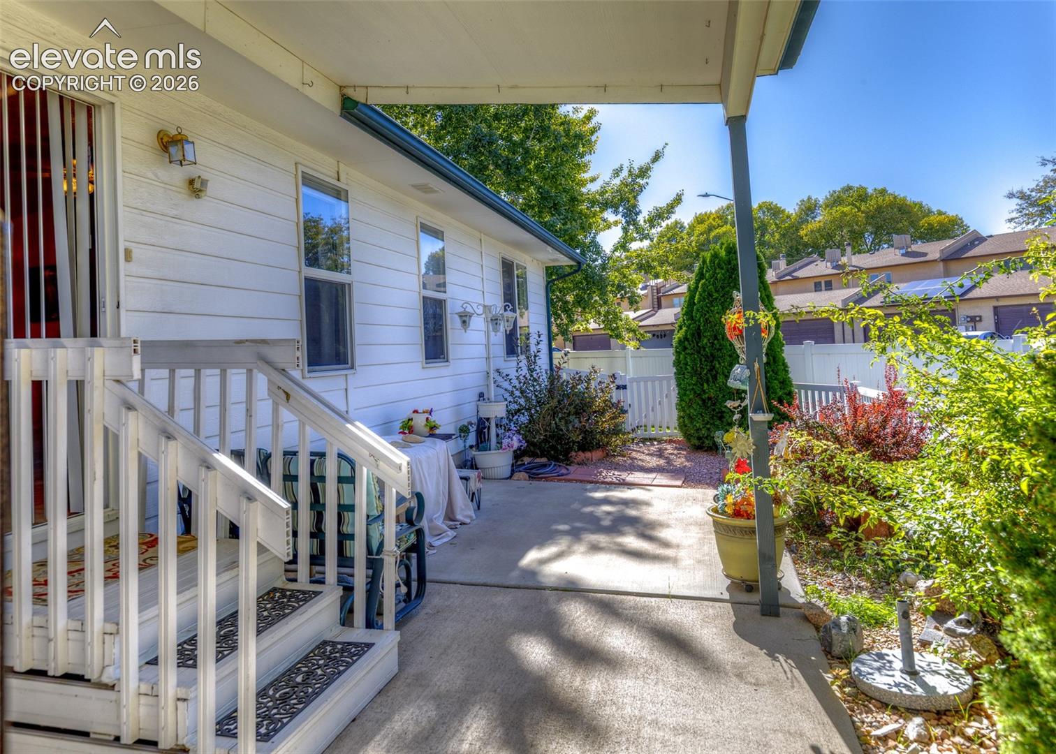 Covered concrete patio featuring white exterior siding, wooden staircase with railings, and white vinyl perimeter fencing