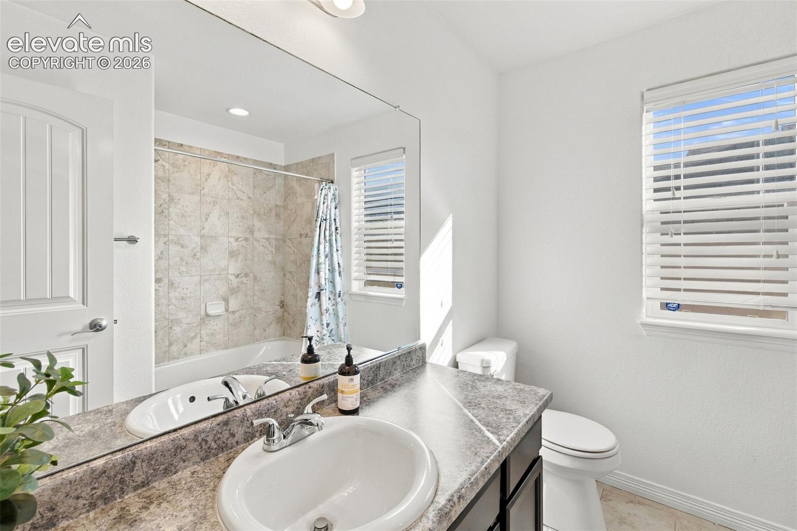 Bathroom featuring a stone-finish countertop, dual-basin vanity, oversized wall-mounted mirror, and a tiled shower surround with tub combination