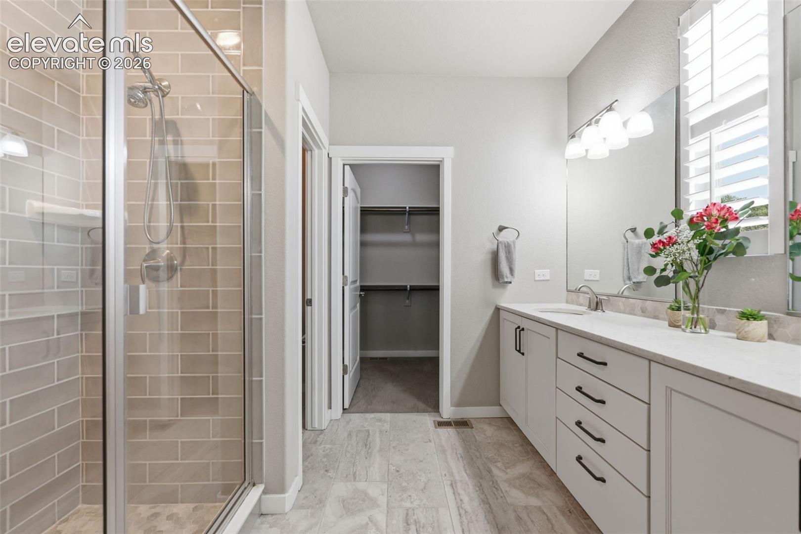 Spacious bathroom featuring a glass-enclosed shower with subway tile, a dual-sink vanity with white stone countertops, and light-colored tile flooring.