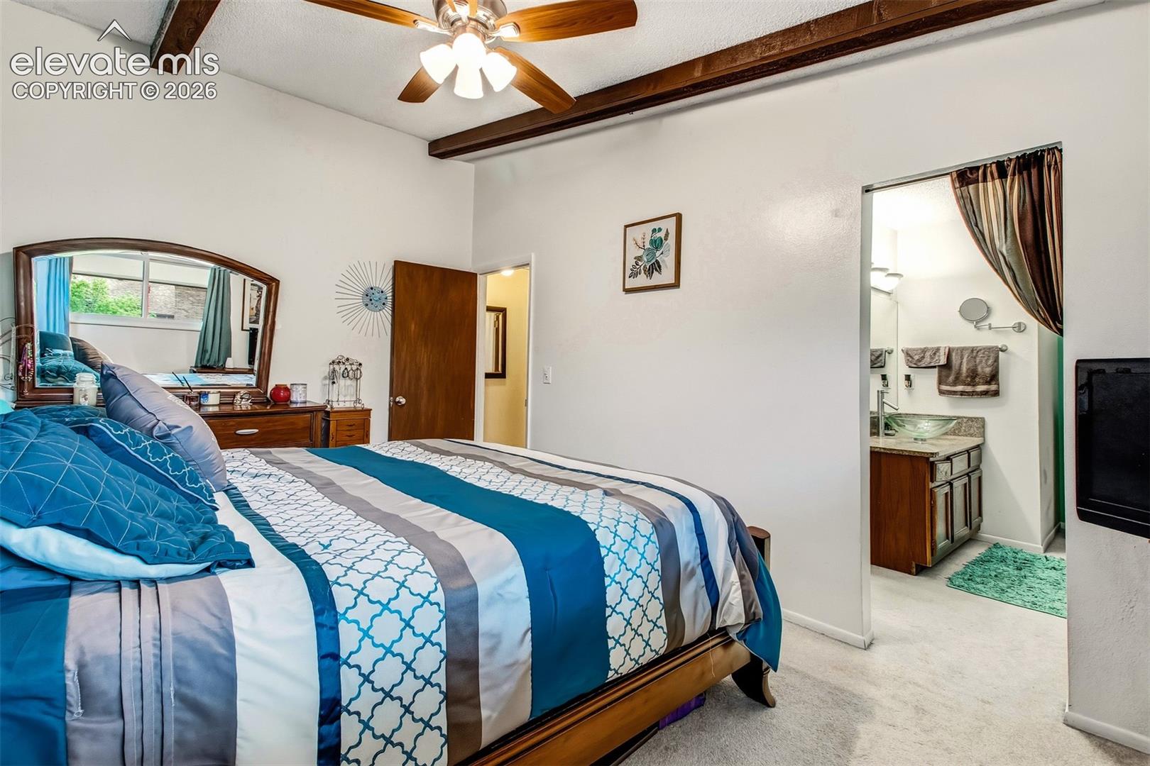 Exposed wood ceiling beams, ceiling fan with light fixture, and an en-suite bathroom featuring a wood-finish vanity and glass vessel sink