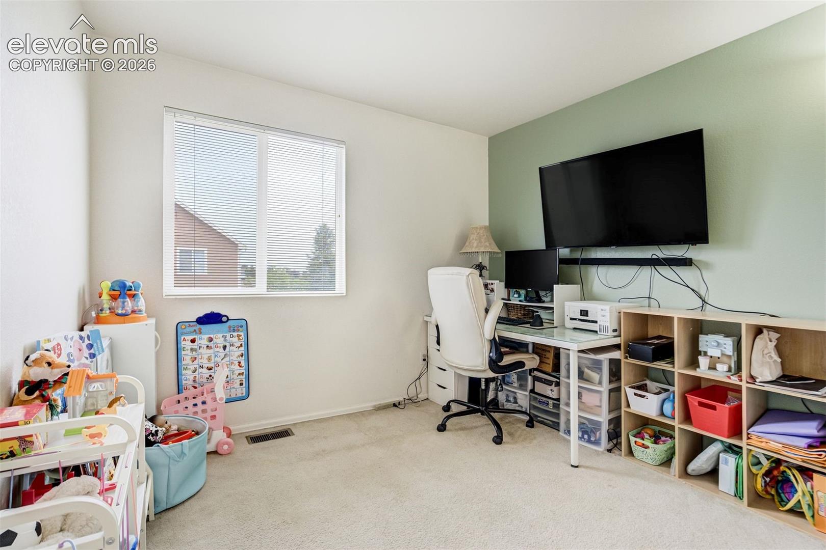 Wall-mounted television with soundbar, modern desk with a glass top, built-in shelving, carpeted flooring, and a large window with horizontal blinds