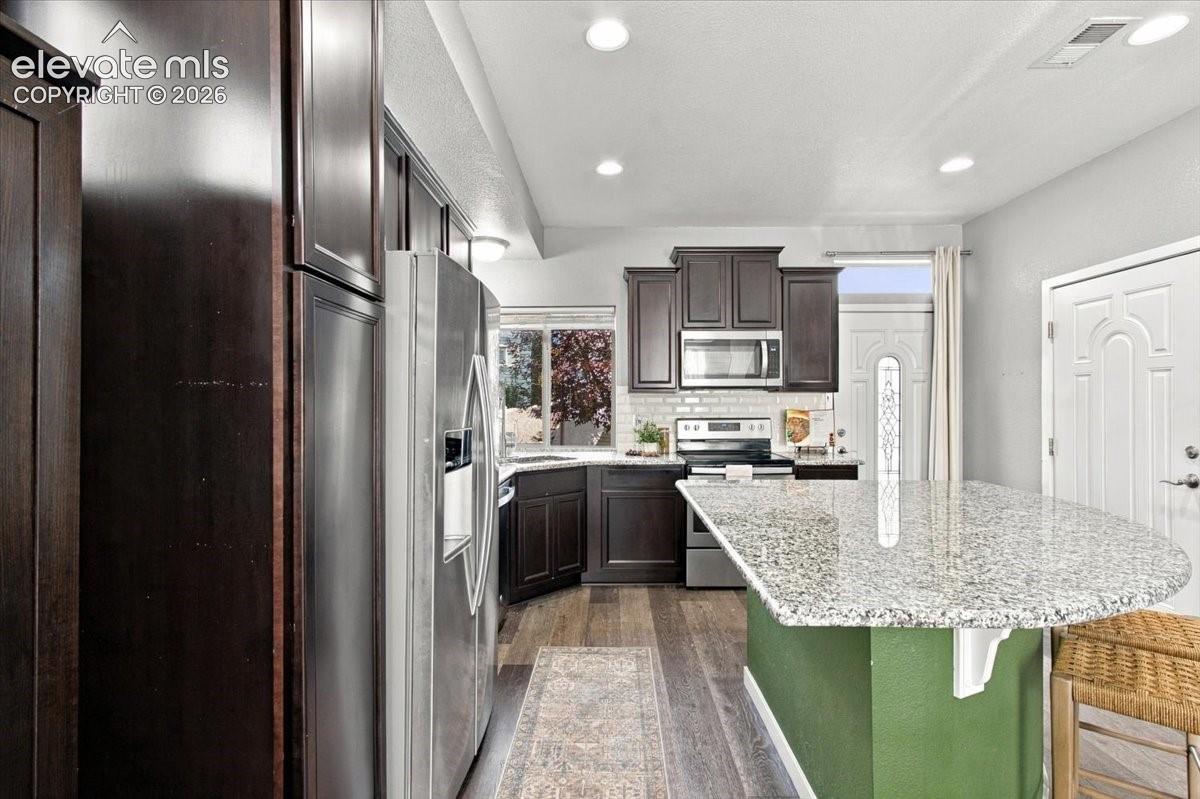 Kitchen featuring dark wood-finish cabinetry, stainless steel appliances, a tile backsplash, wood-finish flooring, and a stone countertop island