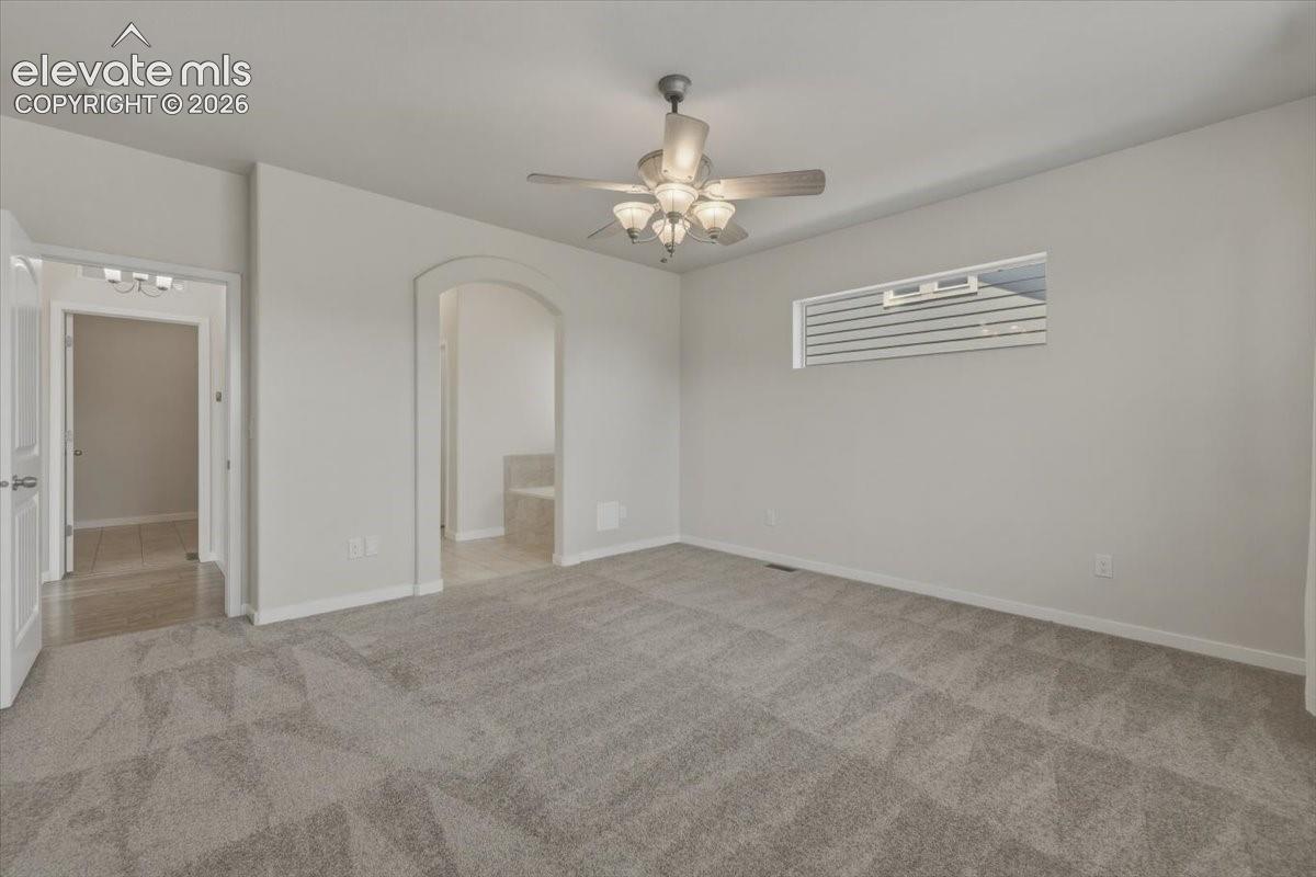 Spacious carpeted room featuring a ceiling fan with integrated lighting, a horizontal window, and an arched entryway leading to a tiled area