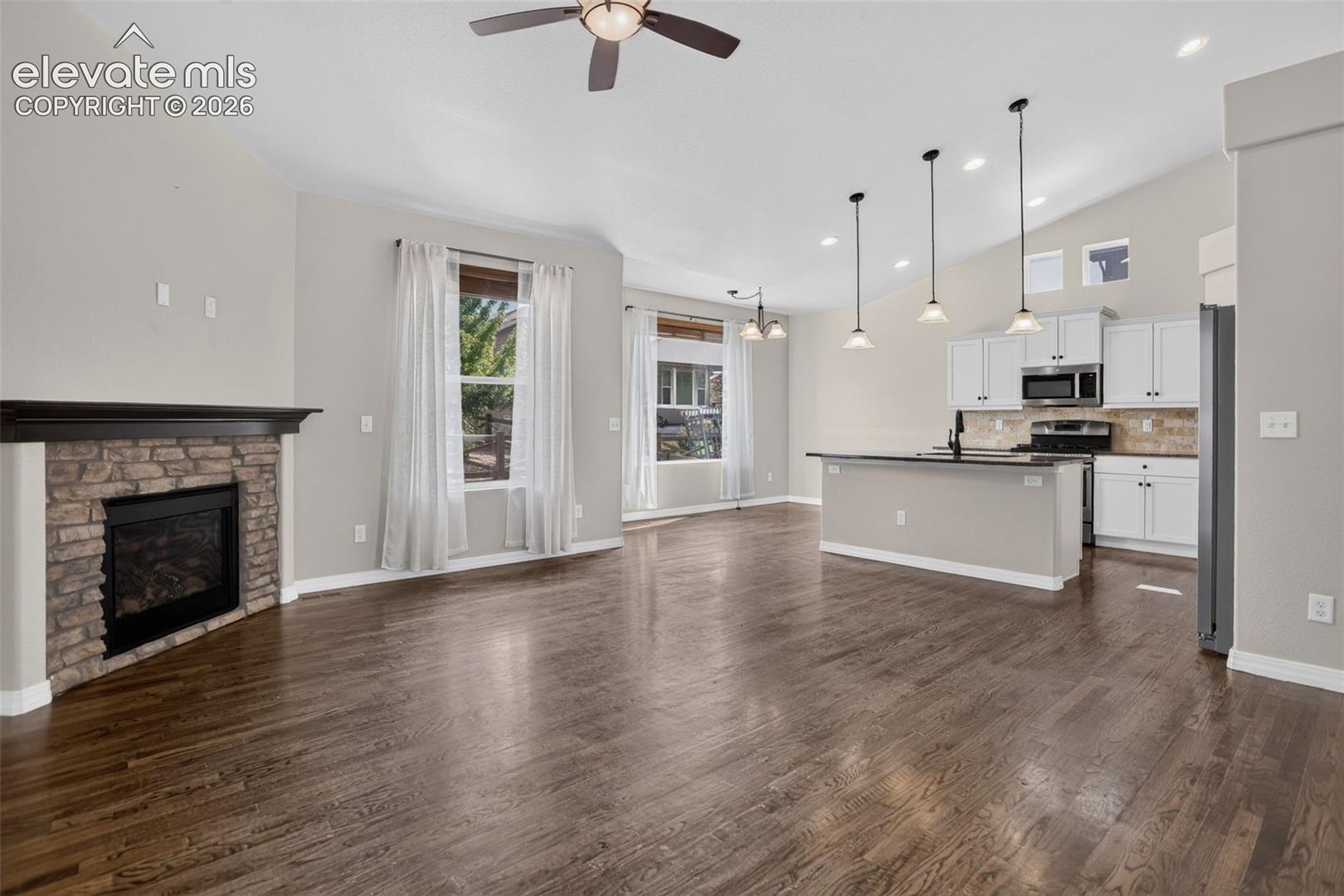 Vaulted ceiling with a ceiling fan, wood-finish flooring, stone-clad fireplace, kitchen island with counter seating, and pendant lighting