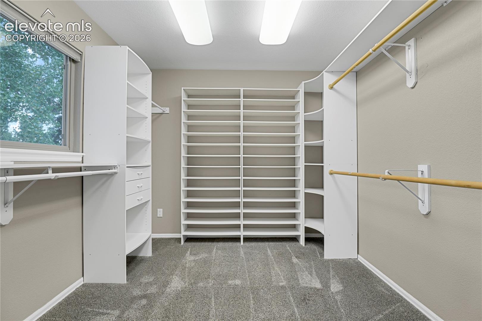 Walk-in closet featuring extensive built-in white shelving, multi-level hanging rods, integrated drawer storage, and windowed natural lighting