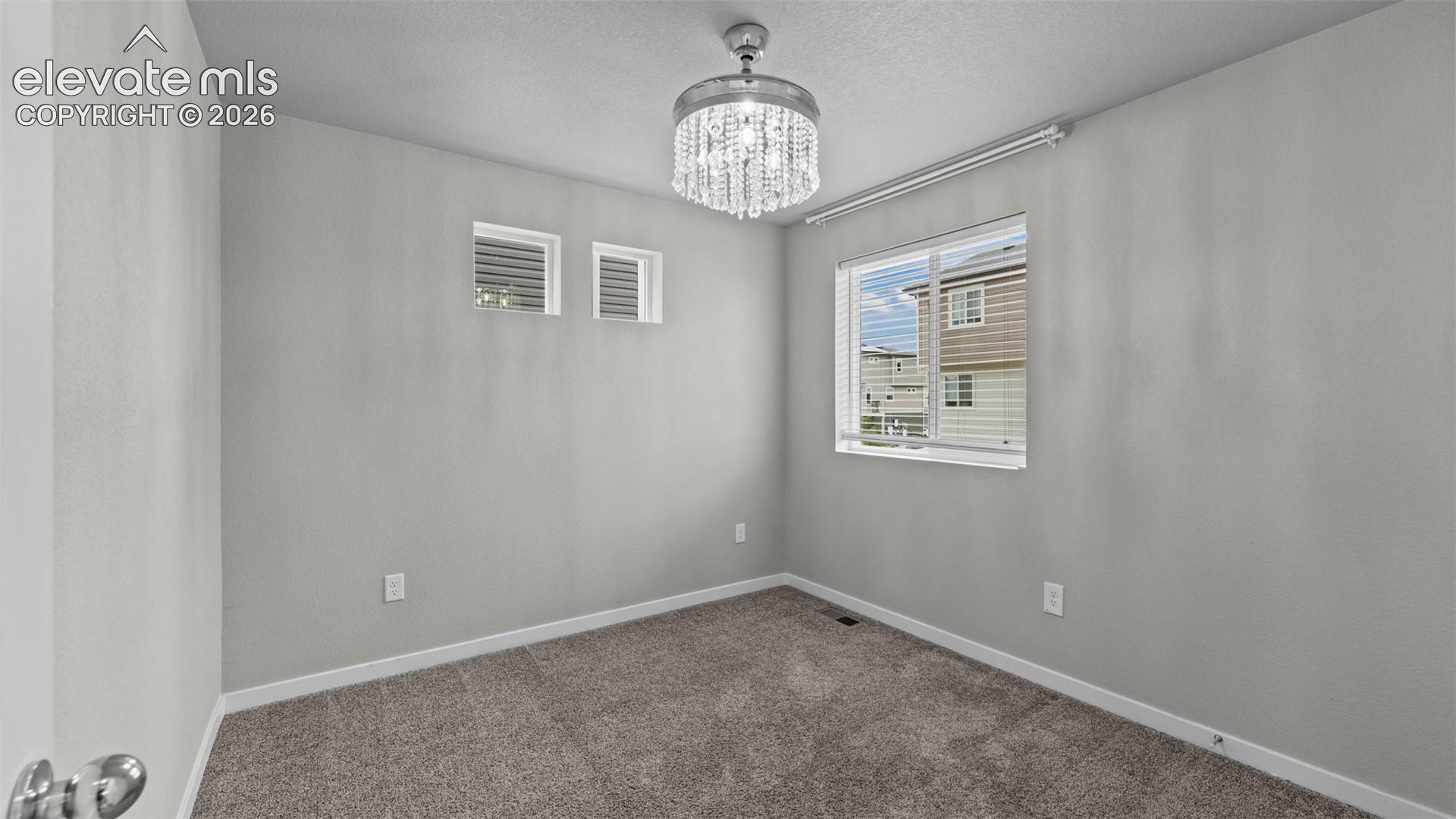 Neutral-toned interior space featuring wall-to-wall carpeting, a crystal-accented ceiling light fixture, dual high-set windows, and a rectangular window with horizontal blinds