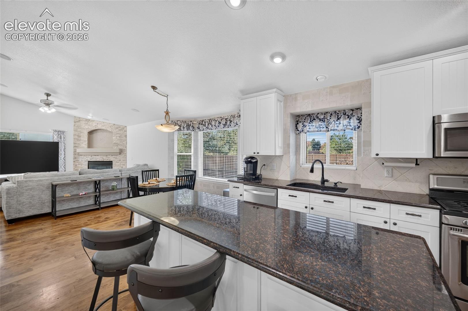 Kitchen featuring white shaker-style cabinetry, dark stone countertops, stainless steel appliances, and wood-finish flooring, complemented by a stone-faced fireplace in the common area