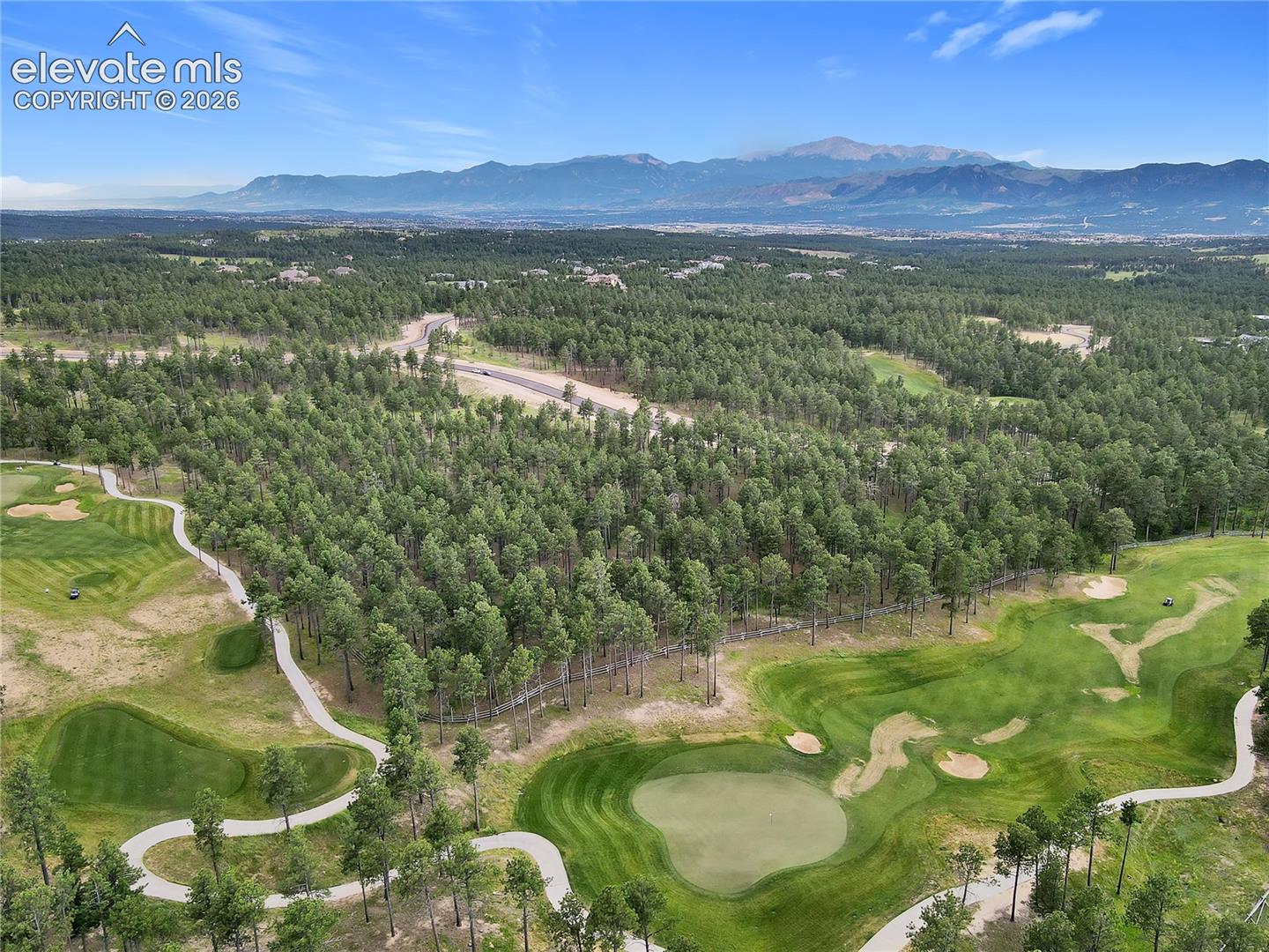 Aerial perspective of a golf course site featuring manicured fairways, extensive wooded areas with mature pine trees, paved cart paths, and mountain peaks in the distance