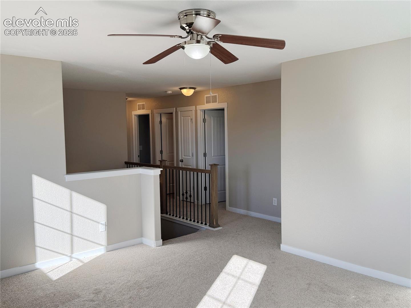 Neutral-toned interior space featuring a five-blade ceiling fan with integrated lighting, carpeted flooring, and a half-wall partition with a metal baluster railing and wood-finish handrail