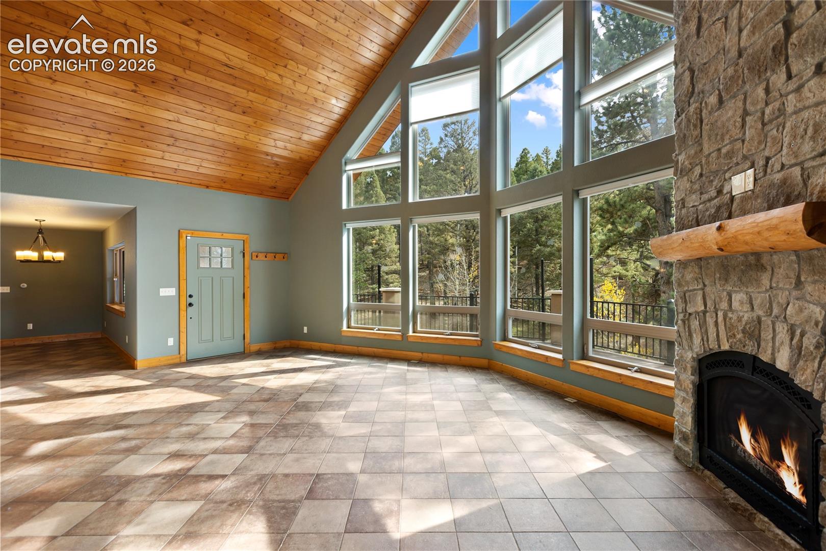Vaulted wood-plank ceiling, floor-to-ceiling multi-pane windows, natural stone fireplace with a thick timber mantel, and large-format ceramic tile flooring