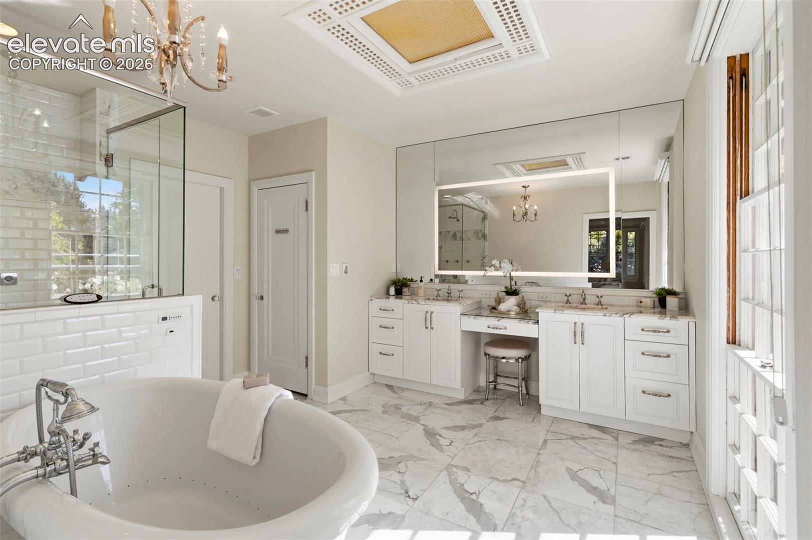 Bathroom featuring a freestanding soaking tub, walk-in glass-enclosed shower with subway tile surround, custom white dual vanity with an integrated makeup station, large wall-to-wall mirror with backlighting, and natural stone flooring