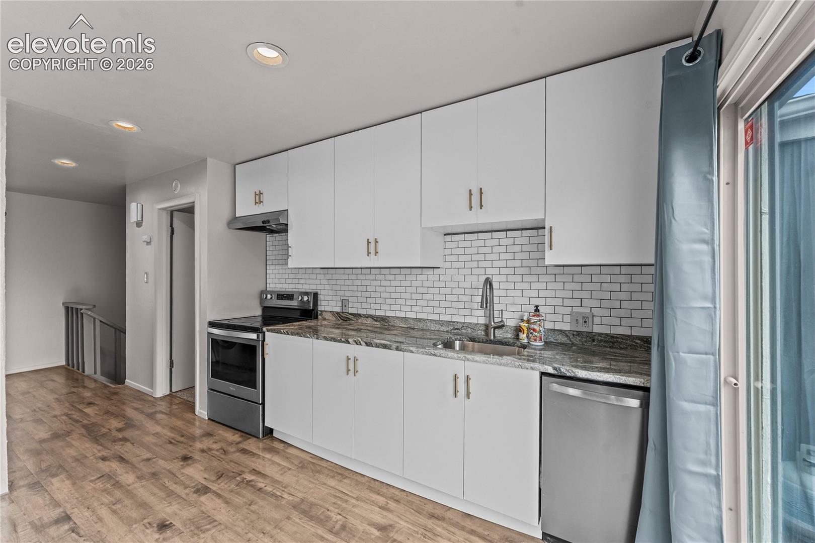 Kitchen featuring white cabinetry with brushed gold hardware, stone countertops, subway tile backsplash, stainless steel appliances, and wood-finish flooring