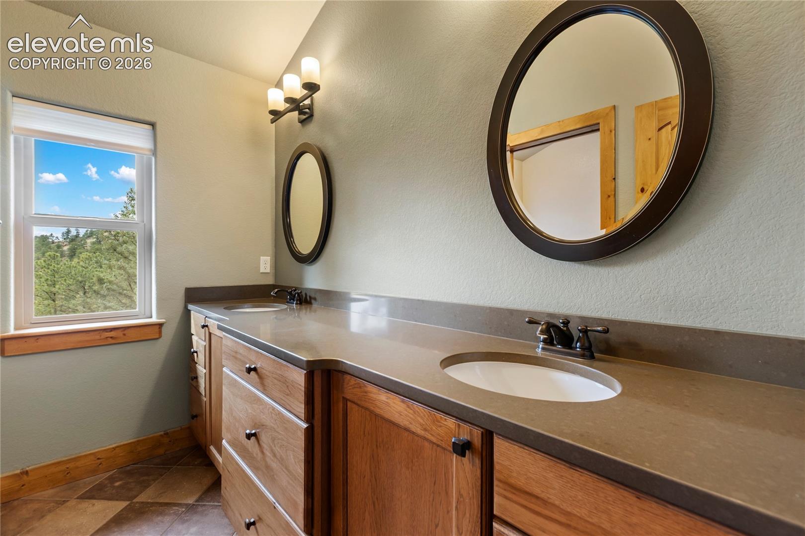 Dual-sink vanity with a solid-surface countertop and undermount basins, wooden cabinetry, dual oval wall-mounted mirrors, and tiled flooring