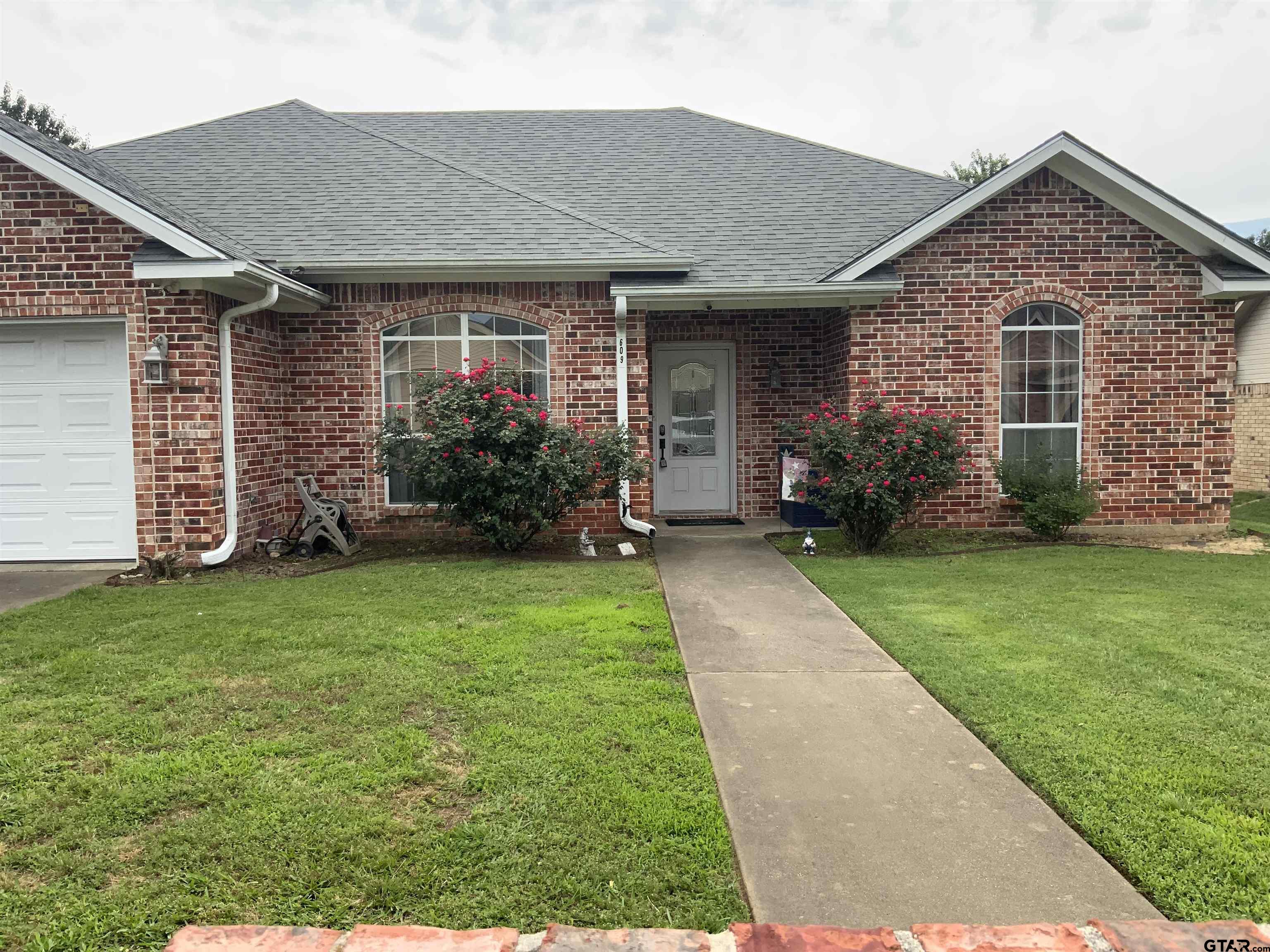 Red brick house with arched windows, a gray roof, flowering roses, and a walkway across the front lawn.