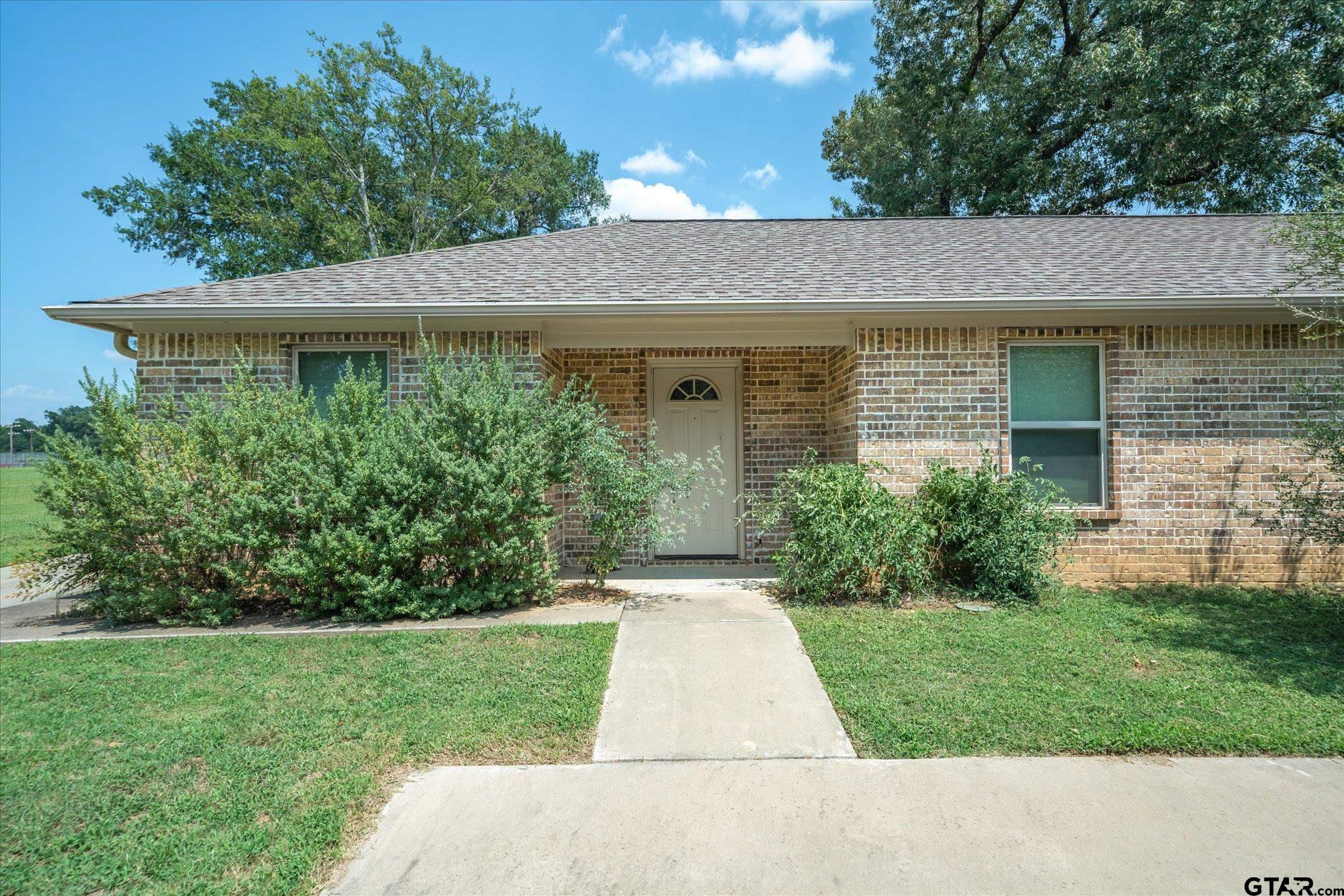 Single-story brick house with a recessed front door, shrubs, and a concrete walkway.