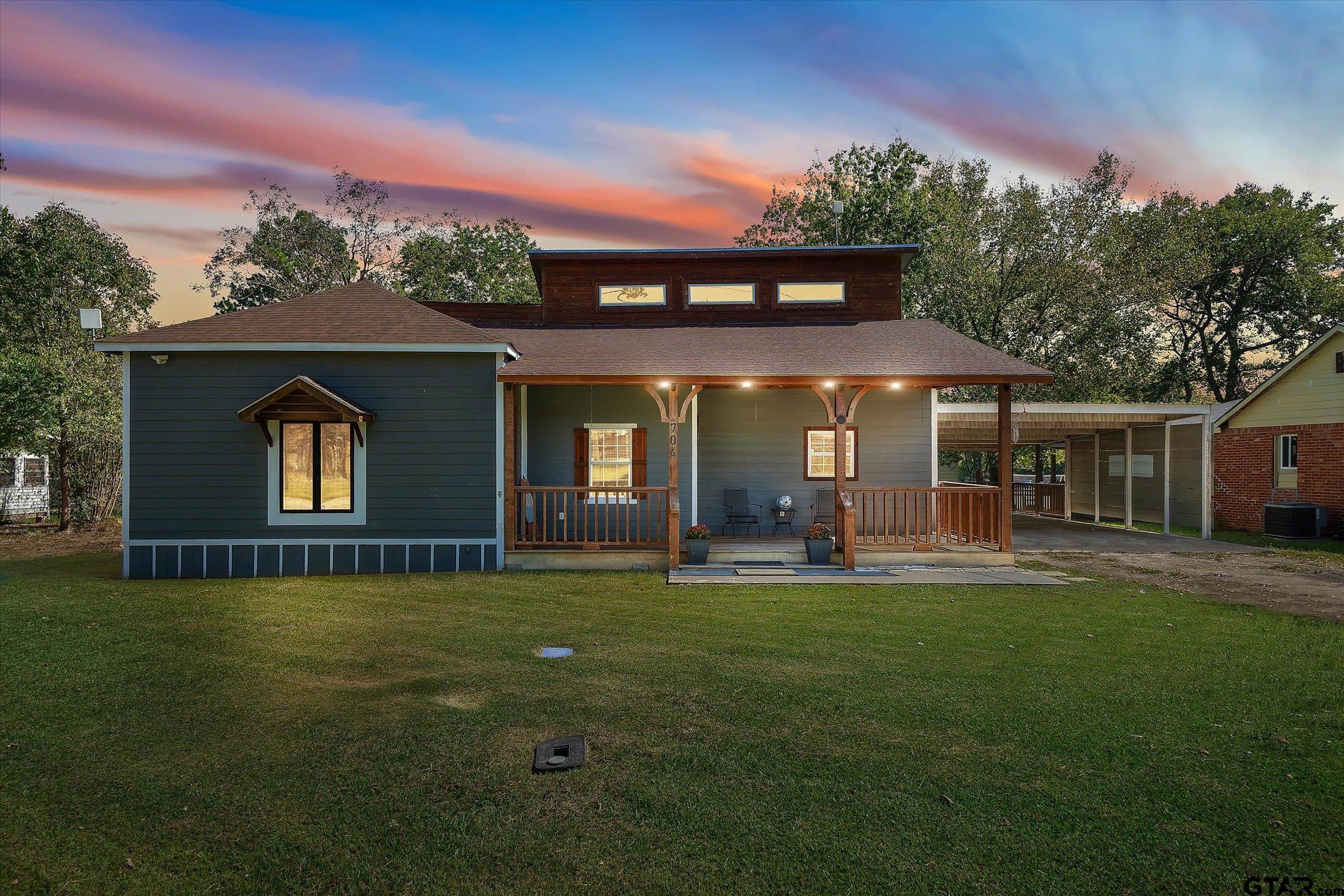 Blue-gray home with a broad timber-covered porch and glowing lights at dusk.