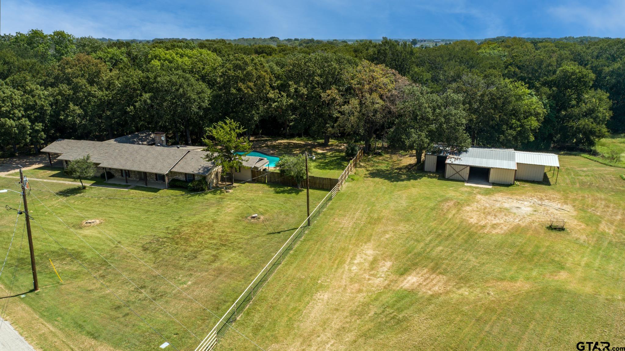 Aerial view of a Wills Point home with a fenced yard, swimming pool, metal outbuilding, and surrounding woods.