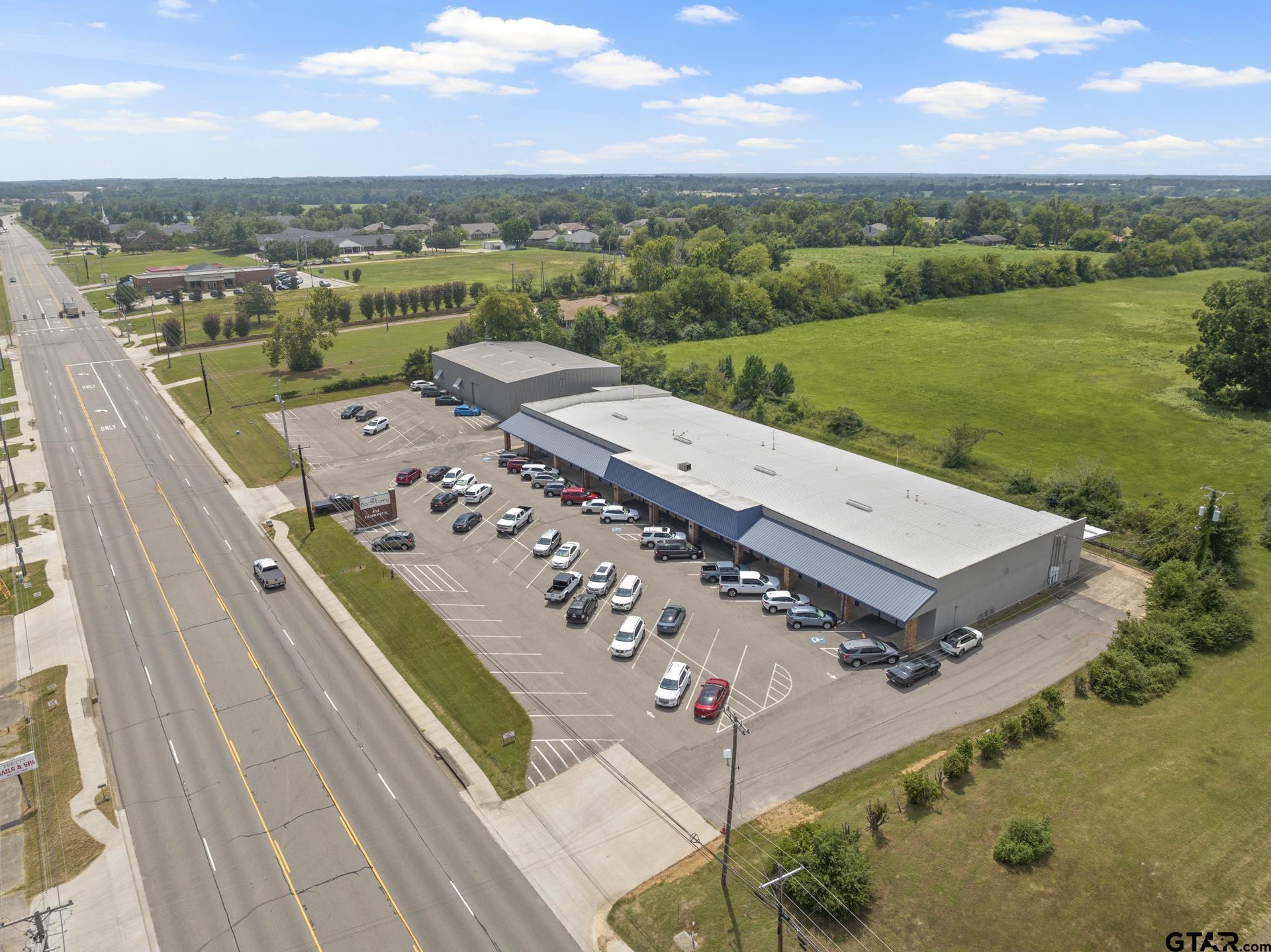 Aerial view of a commercial building with a blue awning and a busy parking lot beside a broad road.