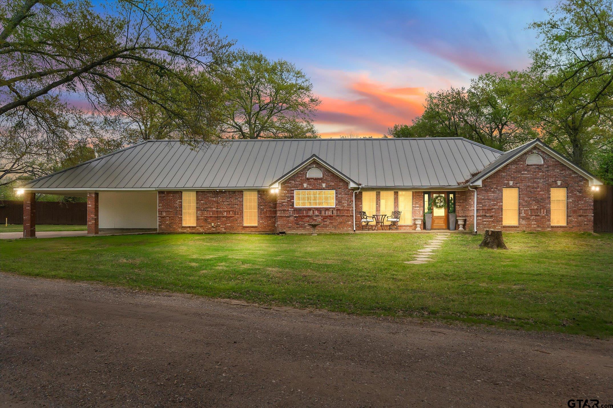 Red brick house with a metal roof and covered carport, warmly lit beneath a colorful dusk sky.