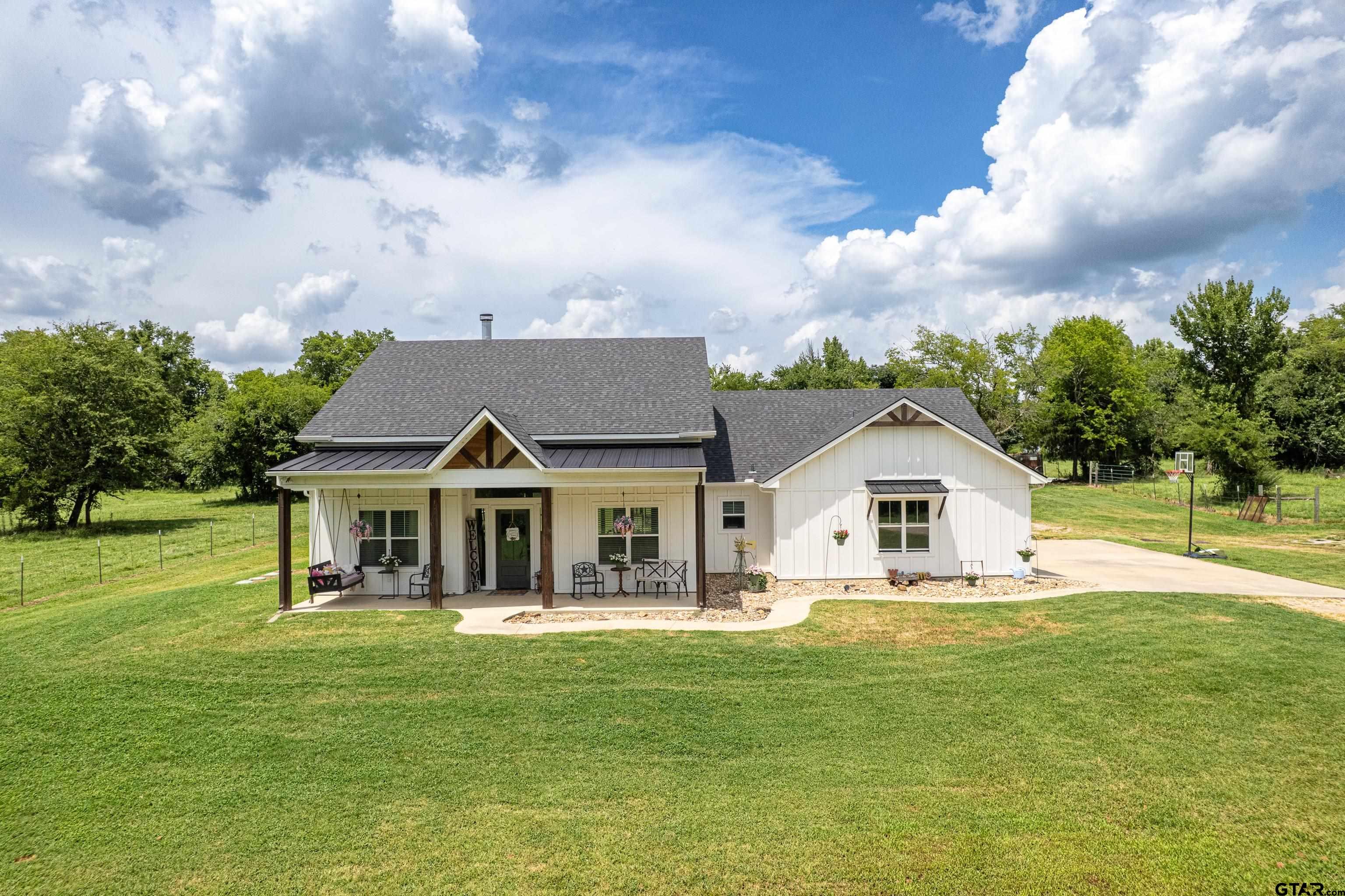 White farmhouse with a covered porch, broad lawn, surrounding trees, and a basketball hoop beside the driveway.
