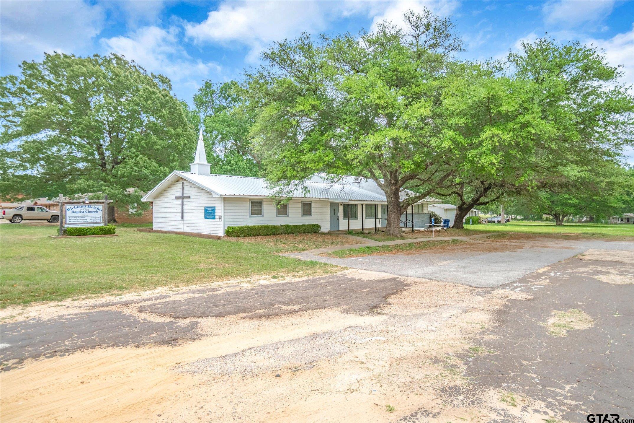 White Baptist church with a steeple beneath large trees beside a paved road.