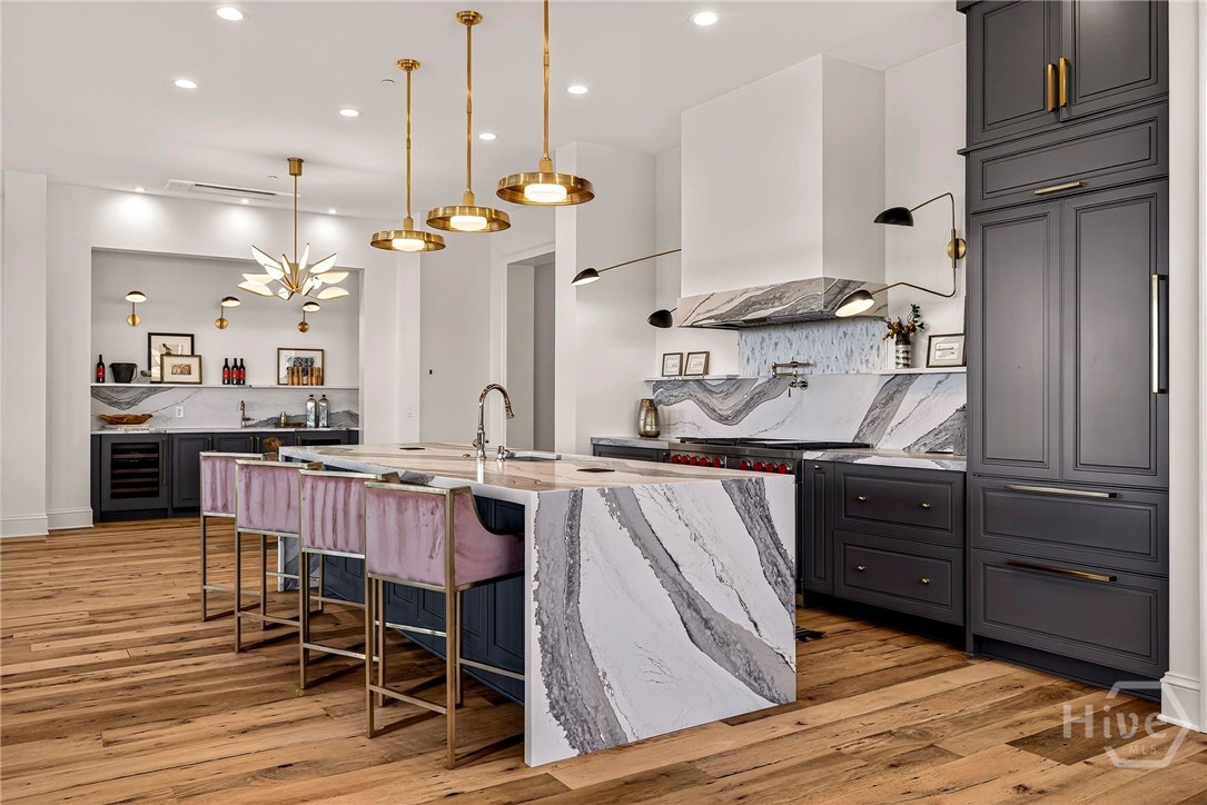 Waterfall island kitchen with integrated appliances. A sculptural waterfall island anchors this luxury kitchen, framed by cohesive marble surfaces and professional-grade appliances. The layout flows into a secondary prep area with wine storage and display shelving, enhancing both utility and elegance. A masterfully planned space for refined culinary living.