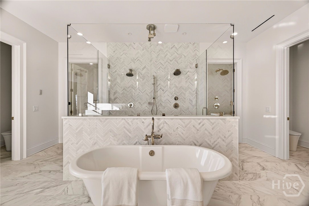 Primary Ensuite enhanced by a sculptural freestanding tub anchors the room, flanked by a symmetrical double-shower setup behind glass partitions. The layout maximizes both privacy and openness, with marble surfaces and recessed lighting reinforcing the spa-like atmosphere. An exceptional primary suite bath designed for restorative living.