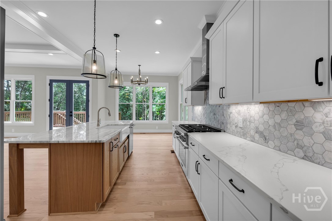 Gourmet kitchen featuring a large central island with a farmhouse sink, light wood-finish flooring, white shaker cabinetry with black hardware, a tiled backsplash, and recessed and pendant lighting