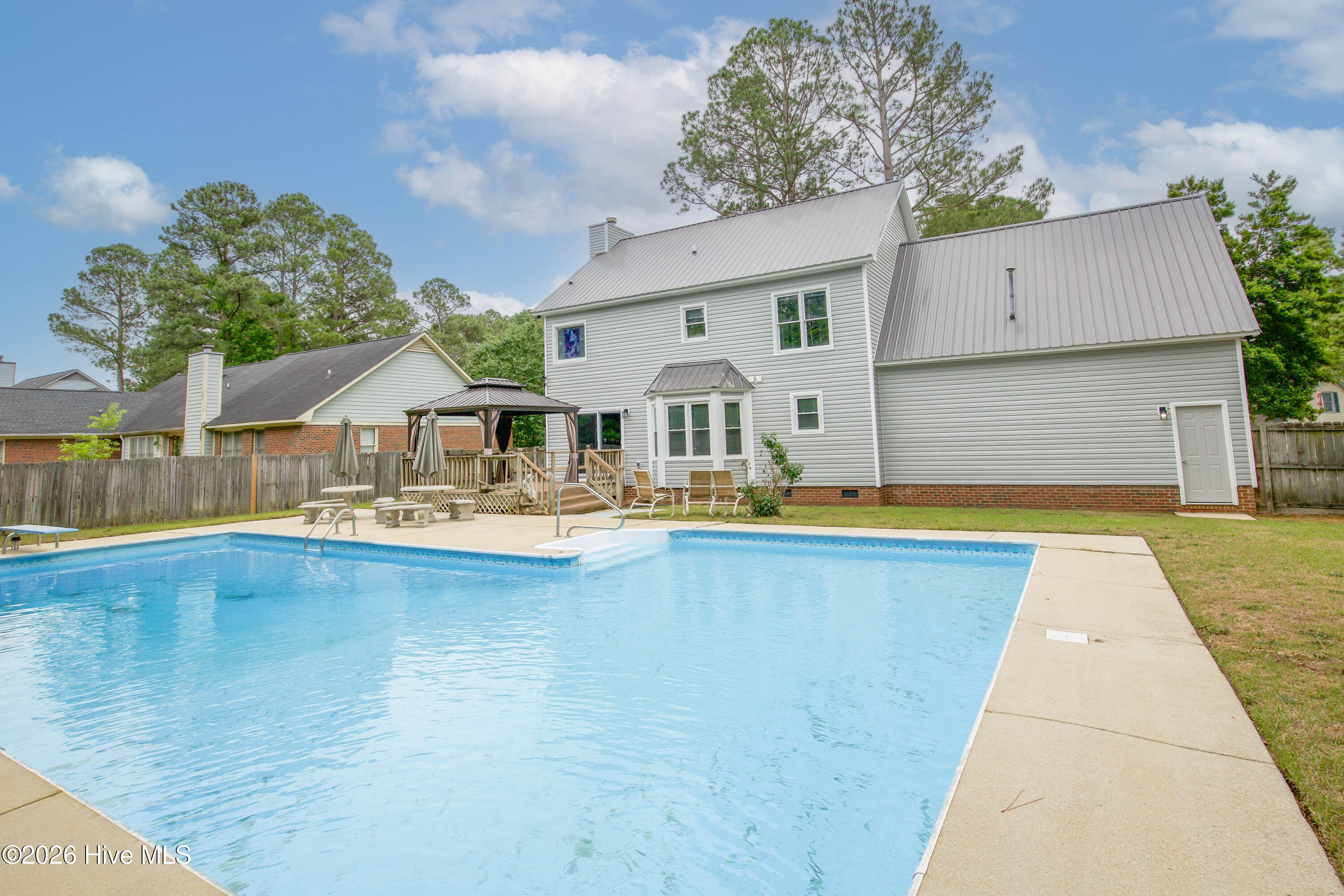 Rear exterior view showcasing the seamless connection between the home and the expansive outdoor entertaining areas. The oversized in-ground pool, spacious patio, elevated gazebo, and fenced backyard create a private retreat ideal for entertaining, relaxing, and enjoying North Carolina summers. Located in the Wells Place community of Fayetteville, NC with no HOA restrictions and convenient access to Fort Bragg, shopping, dining, schools, and medical facilities.