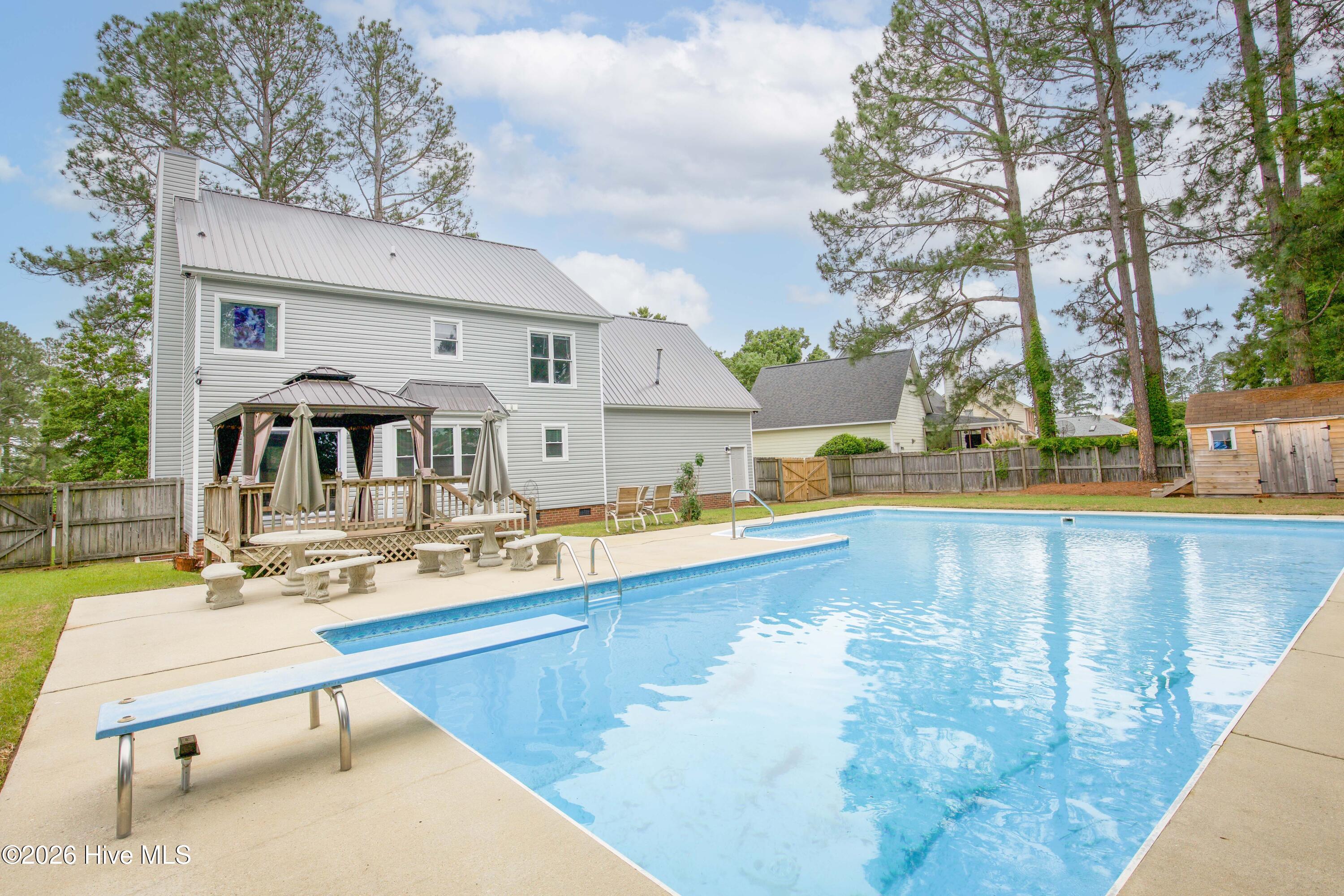 Wide backyard perspective showcasing the impressive size of the in-ground pool, surrounding patio areas, and rear entertaining spaces. The combination of open outdoor living areas, mature trees, and fenced privacy helps create a resort-inspired setting ideal for enjoying North Carolina weather year-round. Conveniently located near Fort Bragg and Raeford Road for an easy commute and access to local amenities.