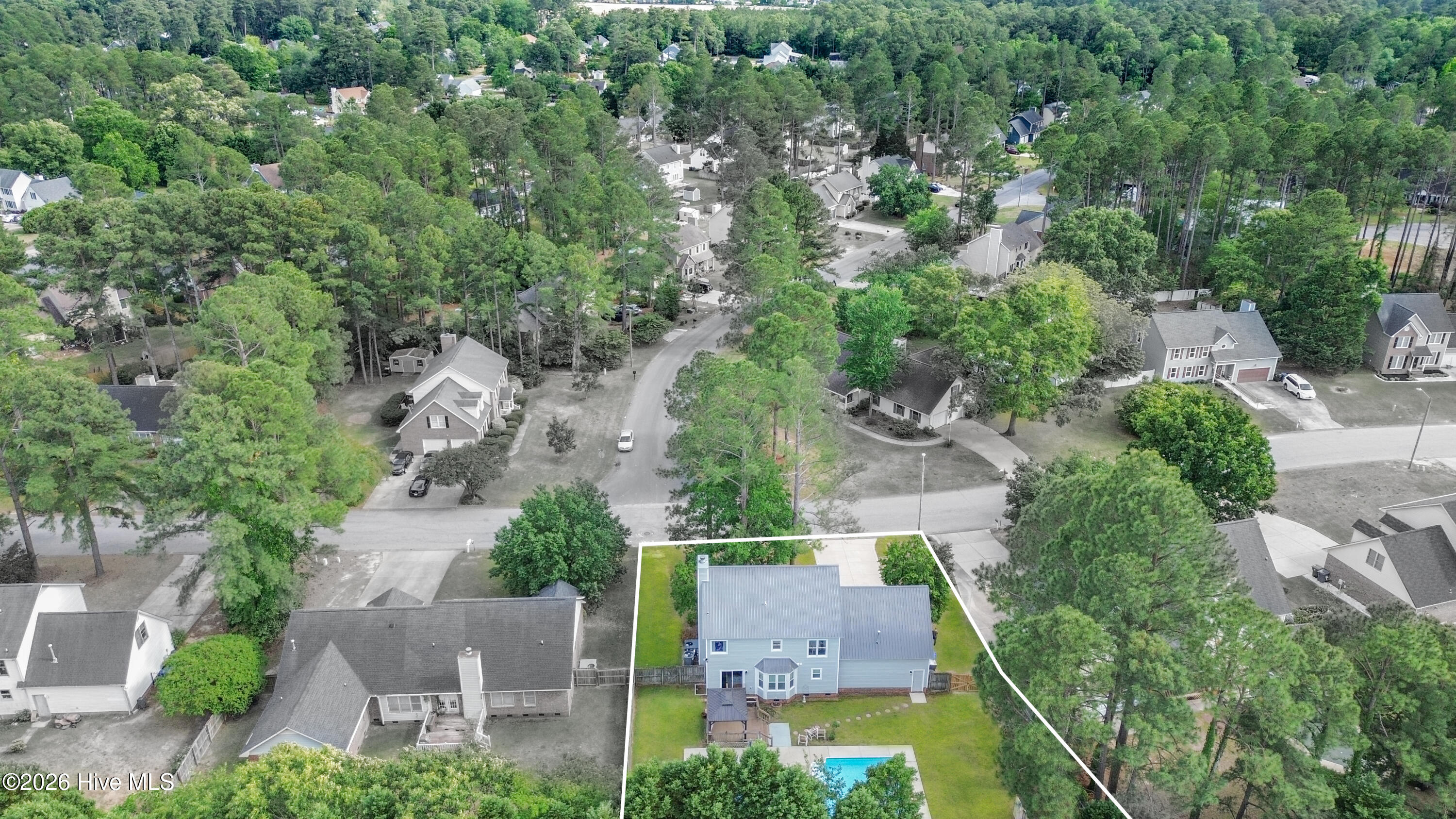 Elevated neighborhood drone view capturing the home’s position within the established Wells Place community surrounded by mature trees and residential privacy. The aerial perspective highlights the backyard retreat, pool area, and convenient Fayetteville location near Fort Bragg and major commuter routes. Aerial imagery may contain virtual enhancements or approximate lot illustrations for marketing purposes only.