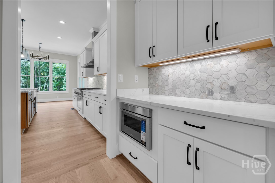 Kitchen featuring bright white cabinetry with black hardware, white countertops, built-in microwave, hexagonal tile backsplash with under-cabinet lighting, and wood-finish flooring