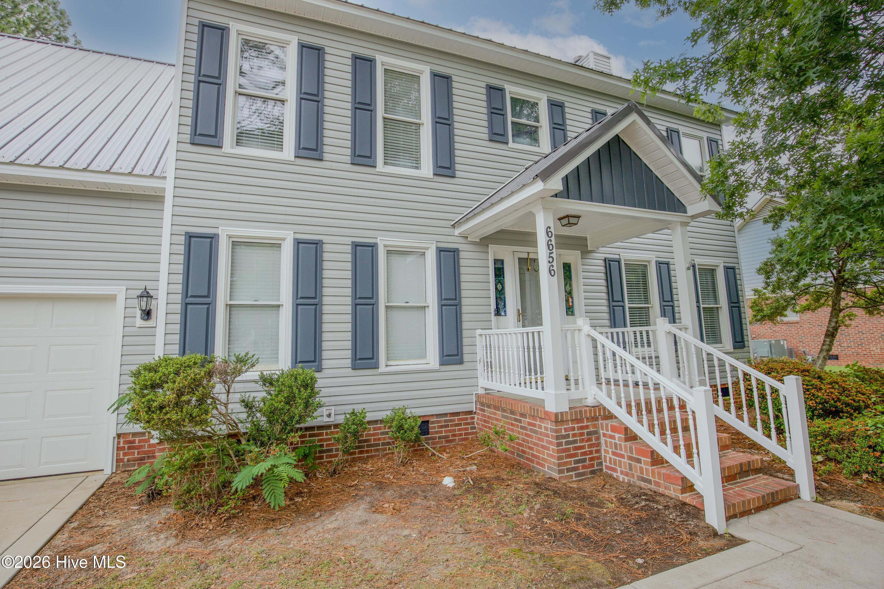 Front exterior angle showcasing the home’s timeless two-story design, covered front porch, oversized two-car garage, and mature landscaping. Located in the established Wells Place subdivision of Fayetteville, NC, this property offers convenient access to Fort Bragg, Raeford Road amenities, medical facilities, and major commuter routes while featuring exceptional outdoor entertaining space and no HOA restrictions.