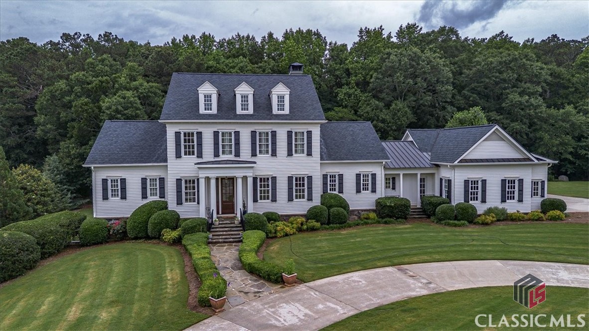 White two-story house with black shutters, three dormers, and a stone walkway, surrounded by mature trees.