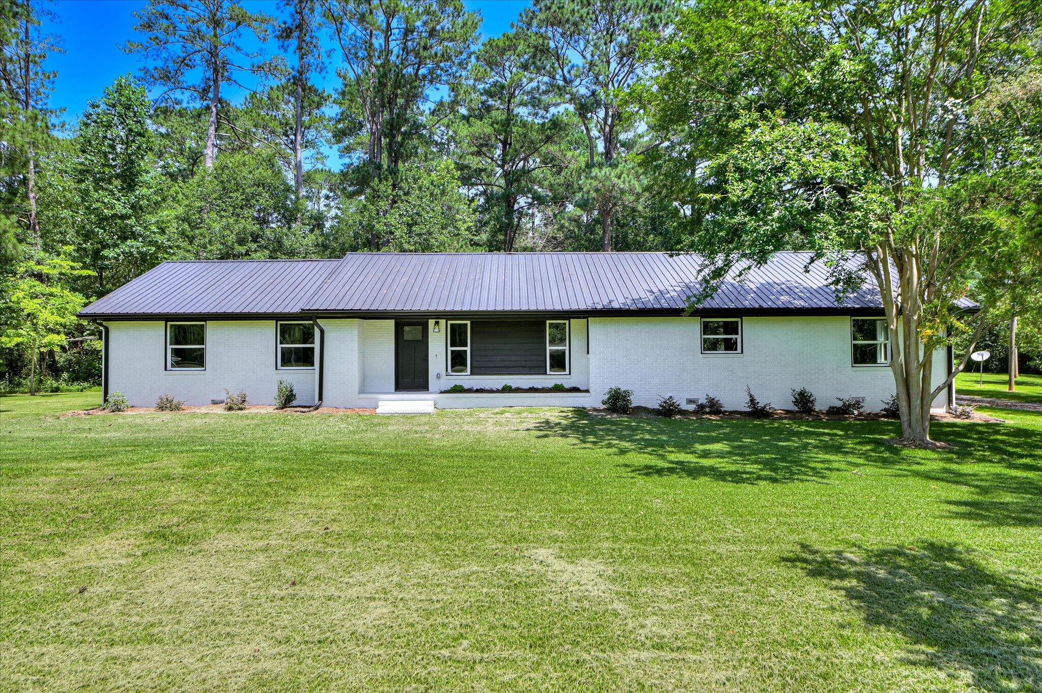 Single-story white brick home with a dark metal roof, broad lawn, and mature trees.