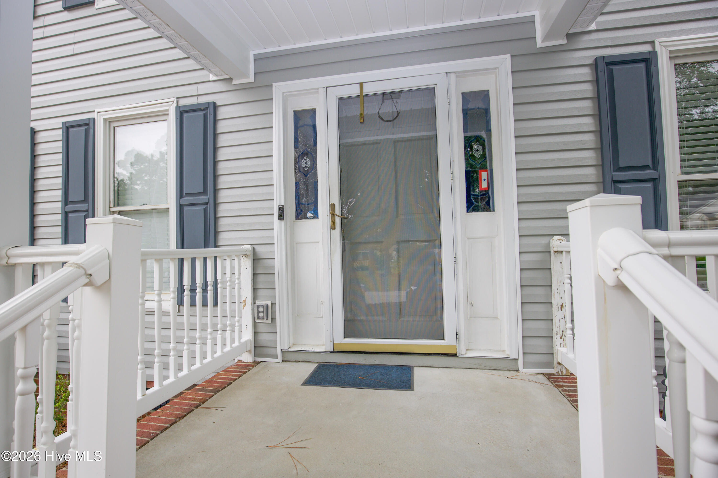 Close-up front entry perspective highlighting the covered porch, upgraded storm door, and welcoming entrance to this move-in ready Wells Place home. The thoughtfully maintained exterior and established landscaping create a warm first impression while reflecting the care and updates completed throughout the property.